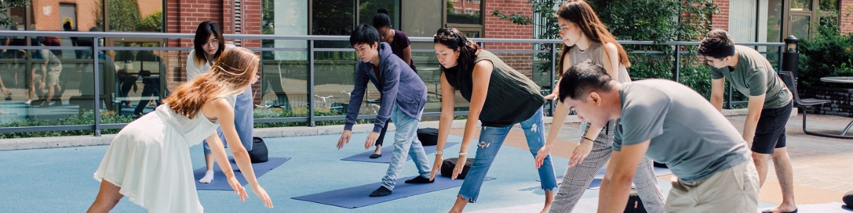 A group of diverse young adults doing mindful stretch exercises on yoga mats outside of an orange brick building in an urban park. They are standing and stretching forwards toward their feet, bending slightly. It is a beautiful sunny day and very bright