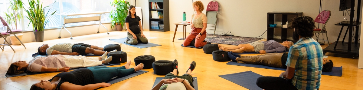 A group of diverse people in a circle deeply meditating on yoga mats inside a very cozy, warm and soothing room. Six people are lying down on their backs and three are sitting comfortably on their knees, including a pregnant fair skinned woman.