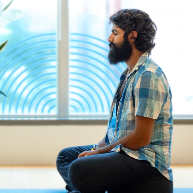 South Asian man with beard wearing a blue plaid shirt meditating while sitting crossed legged on a yoga mat