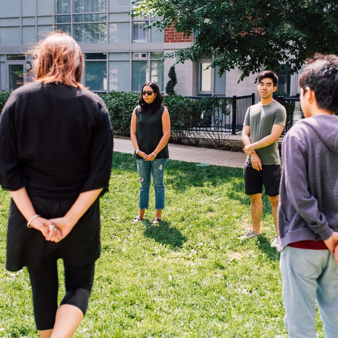 A group of mindful teachers standing wide apart in a large circle in a large green park smiling and listening to a mindful facilitator