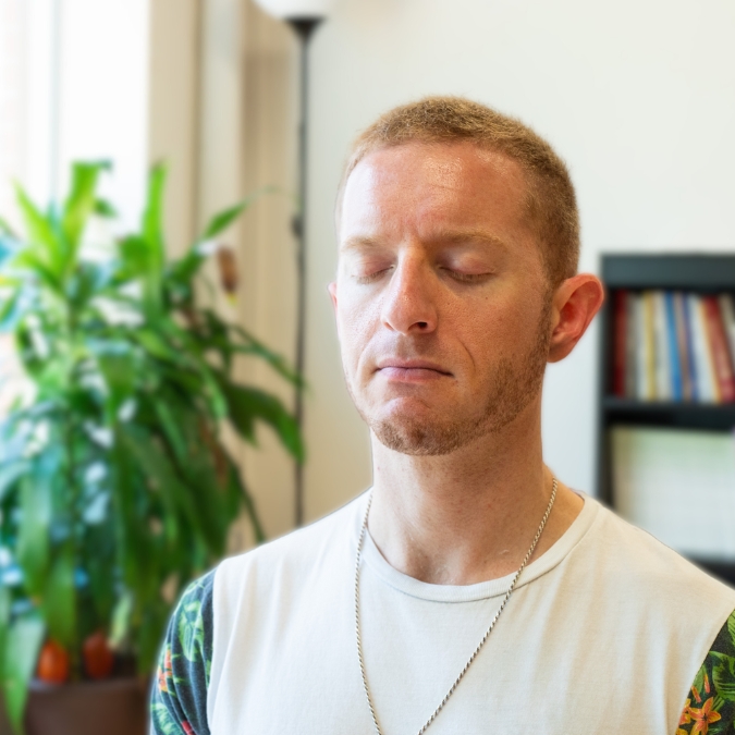 Light skinned man with red hair in deep meditation with eyes closed with plants and bookshelf in the background